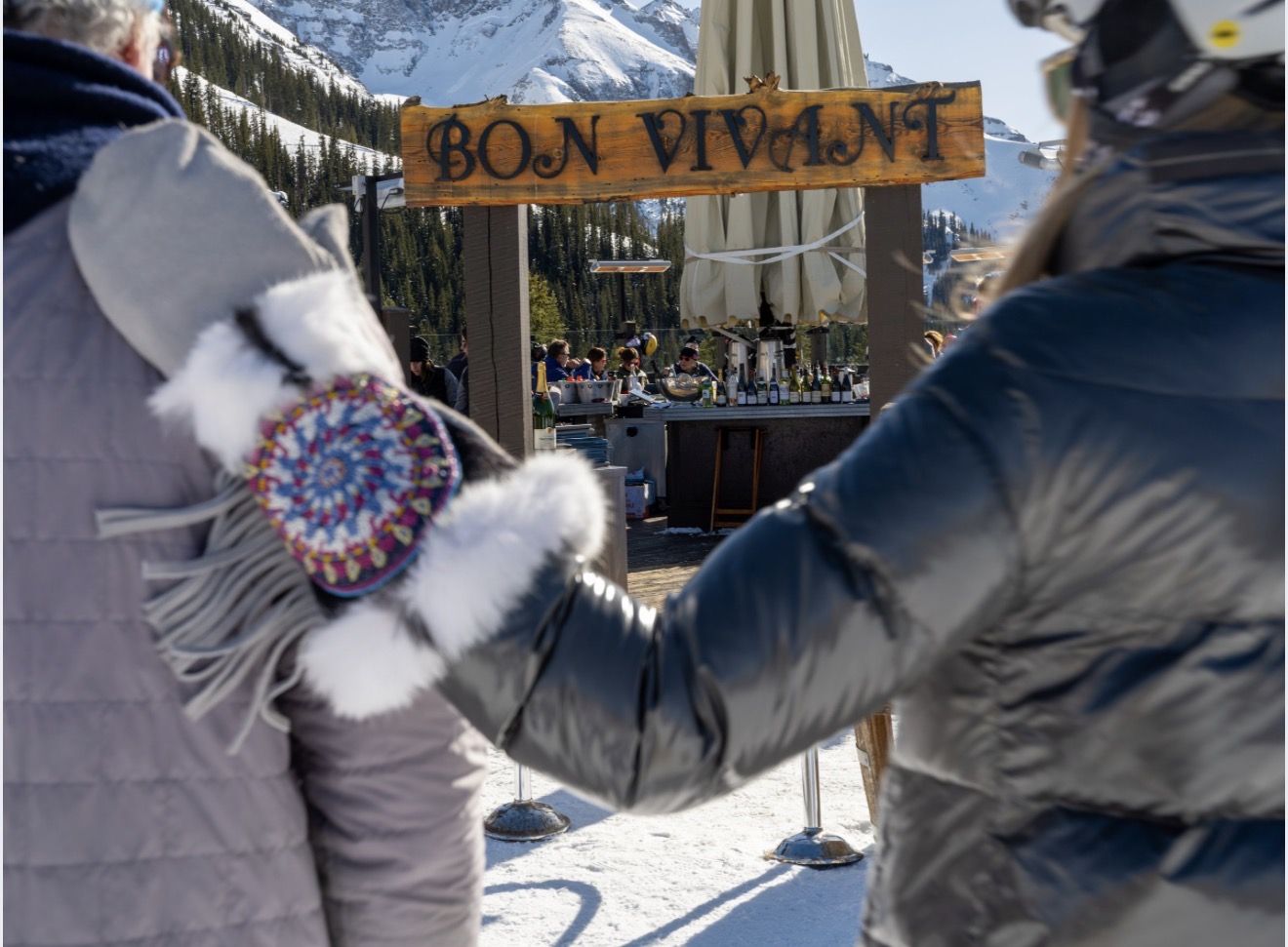 The carved Bon Vivant sign above the outdoor bar at the top of Lift 5, skiers walking in