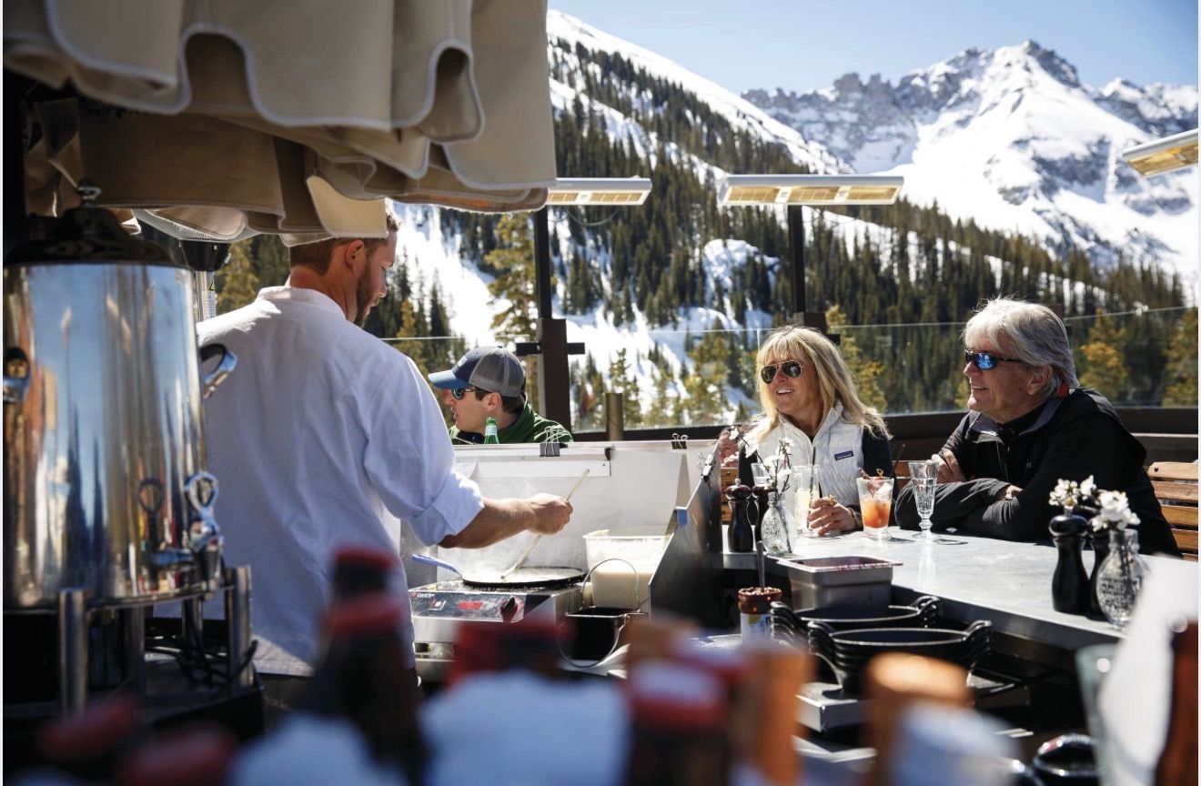 A chef cooking at the counter on the Bon Vivant deck, guests seated at the bar with the mountains behind