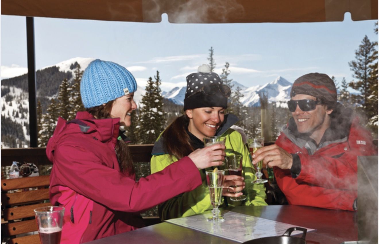Three friends toasting with champagne under the heated umbrella at Bon Vivant, snowy peaks beyond